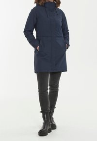 Laivastonsininen vedenpitävä parka, jossa on huppu, vetoketju ja napit, kaksi etutaskua ja hieman kapeneva istuvuus. Valmistettu sileästä ja kestävästä materiaalista.