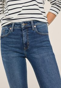 Jeans de mezclilla azules de cintura alta, con diseño de cinco bolsillos, ligero desgaste y accesorios en color cobre, combinados con una camiseta de rayas de mangas largas.