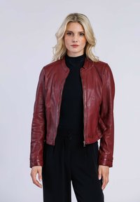 Veste en cuir rouge avec un col montant, fermeture éclair et coupe ajustée. Texture lisse et manches longues, associée à un haut noir.