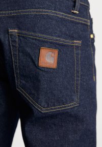 Vaqueros de mezclilla azul oscuro con costuras amarillas y un parche de cuero de la marca Carhartt en el bolsillo trasero.