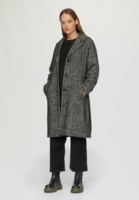 Szary płaszcz w wzór houndstooth z klapami, bocznymi kieszeniami i guzikami, w zestawieniu z czarnymi spodniami i masywnymi czarnymi botkami.