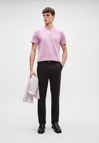 Lys rosa bomulls T-skjorte med en liten logo, kombinert med mørke bukser. Holder en gråstripete skjorte. Svarte sko, stående mot en grå bakgrunn.