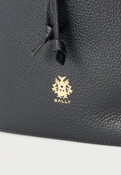 Μαύρη δερμάτινη επιφάνεια με υφή, με μαύρη δερμάτινη γραβάτα και χρυσό λογότυπο Bally που περιλαμβάνει θυρεό και κείμενο.