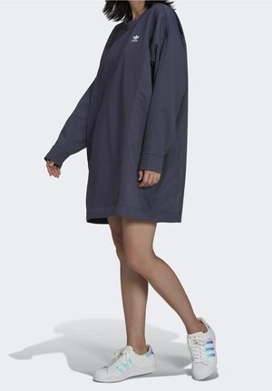 Kvinde iført en oversized mørk Adidas sweatshirt-kjole med hvide Adidas sneakers med irisstriber.