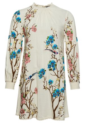 Femme portant une robe fleurie crème à manches longues avec des oiseaux, associée à des chaussettes blanches et des bottines marron, debout contre un mur en bois.