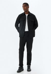Camisa chaqueta oversized de rayas negras con dos bolsillos frontales, combinada con pantalones negros y zapatillas negras, y una simple camiseta blanca.