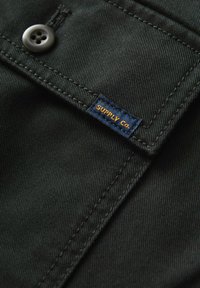 Tissu noir avec une surface lisse et légèrement texturée. Présente une poche cousue avec un bouton sombre et une étiquette marine portant "SUPPLY CO." en orange.