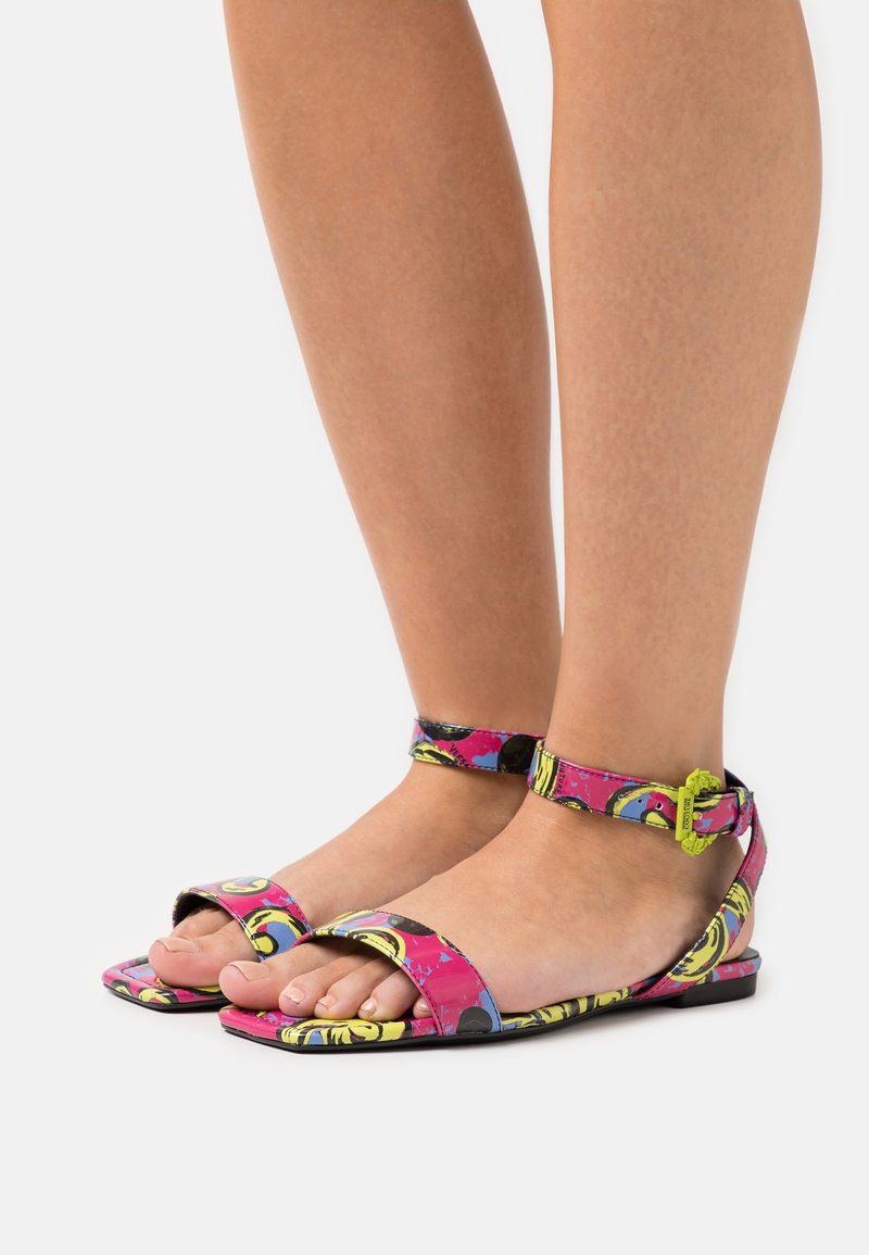 Pieds portant des sandales plates ouvertes aux orteils colorées avec des brides à la cheville et un motif rose, jaune et noir sur fond blanc.