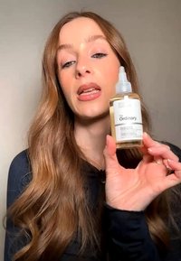 Vrouw met lang golvend haar houdt een klein flesje van The Ordinary huidverzorgingsproduct dicht bij haar gezicht, spreekt of legt uit.