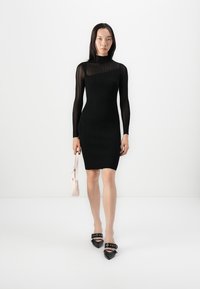 Black ribbed long-sleeve bodycon dress with high neckline. Model holds a small pink handbag, wearing black pointed-toe heels with embellishments.