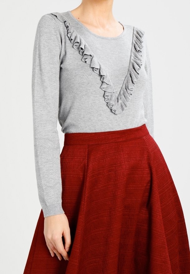 Femme portant un haut à manches longues gris clair avec un bord en V froncé, associé à une jupe rouge texturée taille haute en forme de A.