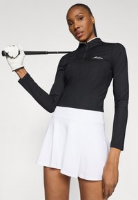 Maglia da golf nera a maniche lunghe con texture a coste e zip a metà, abbinata a una gonna corta bianca svasata. Include un dettaglio con il logo sul petto.