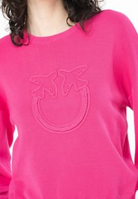 Rosa tröja med ett strukturerat mönster av två fåglar och en flätad cirkeldesign på framsidan. Tillverkad av ett mjukt, elastiskt material.