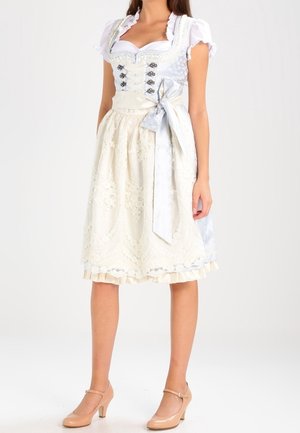 Kobieta ubrana w kremową koronkową sukienkę dirndl z bufkami na rękawach, srebrnymi wiązaniami w talii i cielistymi butami na obcasie Mary Jane, stojąca na białym tle.
