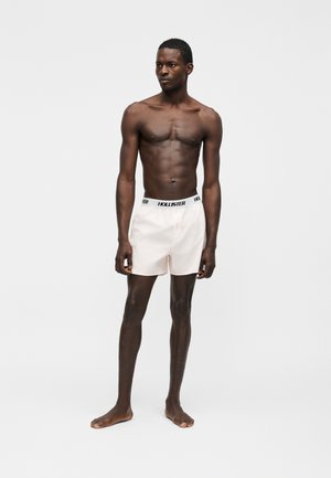 Λεπτός άντρας με σκουρόχρωμο δέρμα, όρθιος και ξυπόλητος, φοράει ανοιχτό ροζ μποξεράκια με ορατή μέση "Hollister", κοιτάζοντας προς τα αριστερά του.