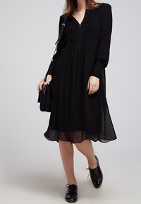 Czarna sukienka z długim rękawem, dekoltem w kształcie litery V, detalami w postaci plisowanego gorsetu, zwiewnym dołem i przezroczystą tkaniną na wierzchu. W połączeniu z czarnymi butami i torebką.