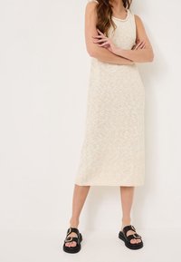 Gebreide crèmekleurige midi-jurk met een aansluitende top, brede bandjes en een losse onderkant. Gecombineerd met zwart sandaaltjes met gouden gespen.