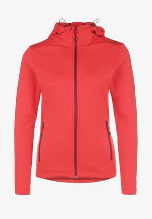 Kobieta idąca ubrana w czerwoną kurtkę na zamek błyskawiczny, szary t-shirt, czarne spodnie i czarne buty trekkingowe na jednolitym jasnym tle.