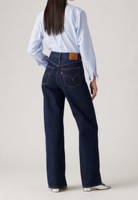 Femme aux longs cheveux noirs portant une chemise bleu clair rentrée dans un jean droit bleu foncé taille haute et des chaussures blanches, vue de dos.