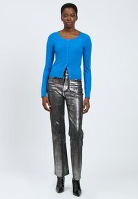 Cárdigan de punto azul con botones en el frente, combinado con pantalones rectos de metal plateado. El modelo lleva botas de tacón negras. Fondo liso.