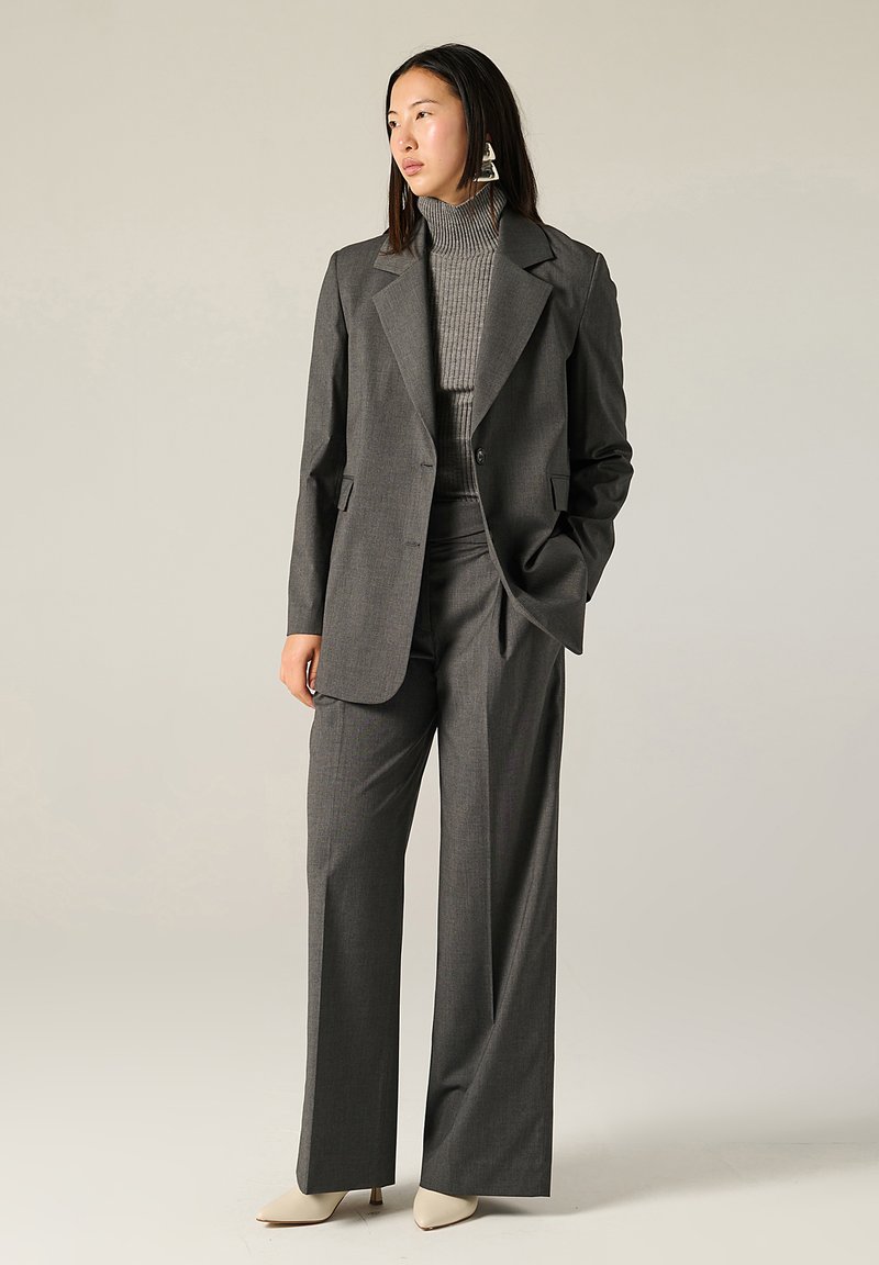 Completo grigio su misura con giacca strutturata a un petto e pantaloni a gamba larga, abbinato a un dolcevita grigio a coste e tacchi di colore crema.