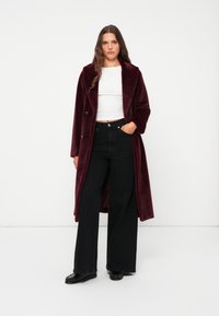 Burgundy long coat made of soft fabric with a notched collar, double-breasted design, worn over a white top and black high-waisted jeans.