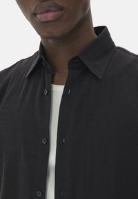 Chemise noire à manches longues avec un tissu texturé, présentant un col classique et une patte de boutonnage avec des coutures et des boutons visibles. Portée par-dessus une chemise blanche.