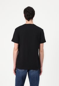 Schwarzes Baumwoll-T-Shirt mit kurzen Ärmeln und rundem Ausschnitt, von hinten präsentiert. Eine blaue Jeans ist auf Taillenhöhe sichtbar.