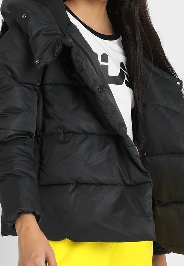 Personne portant une doudoune noire par-dessus un t-shirt blanc graphique et un pantalon jaune vif, visible des épaules aux hanches.