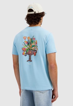 Man wearing light blue t-shirt with colorful floral vase graphic on back and white cap with adjustable strap, standing against plain background.