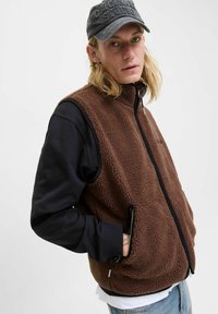 Blonde Person trägt eine schwarze Mütze, eine braune Fleeceweste über einem schwarzen Langarmshirt, mit den Händen in den Vest-Taschen, schaut in die Kamera vor einem weißen Hintergrund.