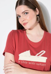 Camiseta de color óxido con mangas cortas y cuello redondo, que presenta un gráfico de una sandalia y el texto "en sintonía con la naturaleza" en blanco.