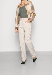 Beige en bruine geruite jas, groene gebreide crop top, lichtbeige stretchbroek met elastische taille en donkergroene gestippelde hakken.