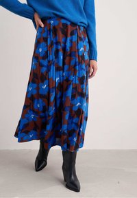Donna che indossa una gonna midi floreale blu con foglie marroni e nere, un maglione blu e stivali con tacco a punta neri, in piedi su un pavimento di cemento.
