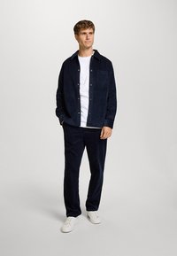 Marineblaue Cord-Hemdenjacke mit durchgehender Knopfleiste und Brusttasche, kombiniert mit passenden Cordhosen. Weißes T-Shirt und Turnschuhe.
