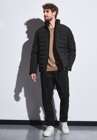 Černá péřová bunda s prošívaným designem, vysokým límcem a předním zipem, nošená přes béžový svetr a spojená s černými kalhotami a bílými teniskami.