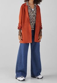 Abrigo de ante en color óxido sobre una camisa de estampado de leopardo, combinado con vaqueros de pierna ancha en azul y zapatillas en blanco y negro, con una capa inferior de red.