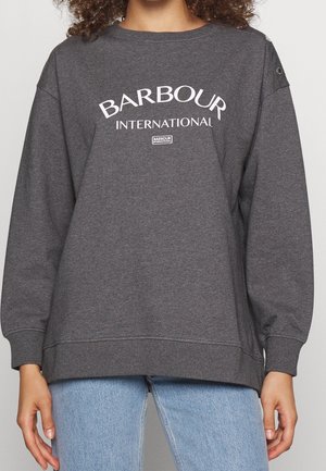 Dark gray Barbour International sweatshirt with white logo text, worn by a person with light blue jeans, cropped at neck and hips.