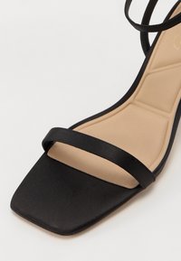 Sandale noire à bout ouvert avec bout carré, une seule bride avant et une semelle intérieure beige rembourrée avec des coutures géométriques.