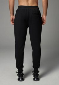 Zwarte sweatpants met een gestructureerde stof, een elastische tailleband en een enkele achterzak, gecombineerd met zwarte sneakers, weergegeven van achteren.