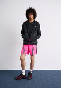 Sort hættetrøje med lynlås og hvidt logo, sort t-shirt, livlige pink shorts med grå detaljer og sorte sportssko med røde detaljer.