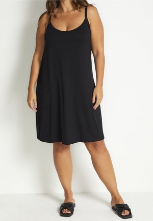 Femme portant une robe noire sans manches jusqu'aux genoux et des sandales noires sans lacets, debout devant un fond blanc uni.