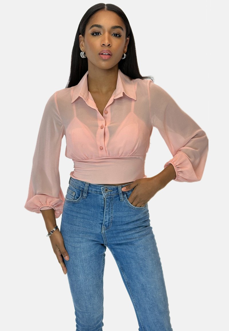 Blusa rosa trasparente con bottoni frontali, design con colletto e maniche a sbuffo. Indossata con jeans blu a vita alta, che mettono in risalto una silhouette aderente.