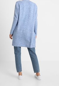 Osoba w długim jasnoniebieskim swetrze, niebieskich dżinsach i białych trampkach, stojąca plecami do aparatu na jasnym, jednolitym tle.