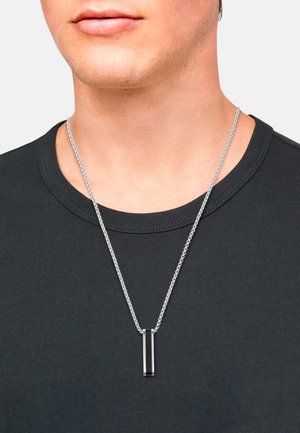 Collier en argent avec un pendentif rectangulaire noir. Le pendentif a une finition lisse, attaché à une chaîne texturée, porté sur une chemise noire.