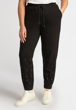 Pantaloni della tuta neri con polsini elasticizzati, vita con cordoncino e accenti in perline argentate lungo le gambe. Abbinati a scarpe da ginnastica bianche.