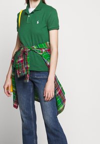 Grön polo skjorta med en logotyp, i kombination med blå jeans och en rutigt flanell skjorta knuten runt midjan i grön, röd och gul. Gul handväska.