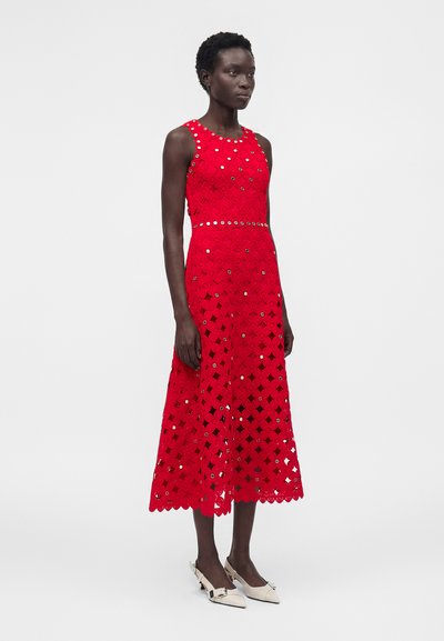 Γυναίκα φορούσε κόκκινο δαντελένιο midi φόρεμα χωρίς μανίκια με καθρεφτιστές διακοσμήσεις και κυματιστό τελείωμα, σε συνδυασμό με λευκές μυτερές γόβες.
