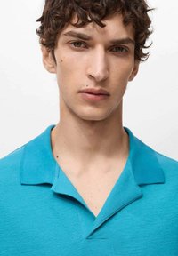 Turkizna polo majica z teksturirano tkanino, z klasičnim ovratnikom in placketom s tremi gumbi, nosi model na belem ozadju.
