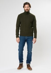 Pulover verde tricotat cu guler înalt, model cu torsade, cu manșete și tiv ribbelate, asortat cu blugi albaștri și cizme maro cu șireturi.